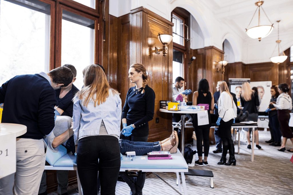 Delegates working on reclined models at treatment couches in a large training room, with gloved trainers supervising