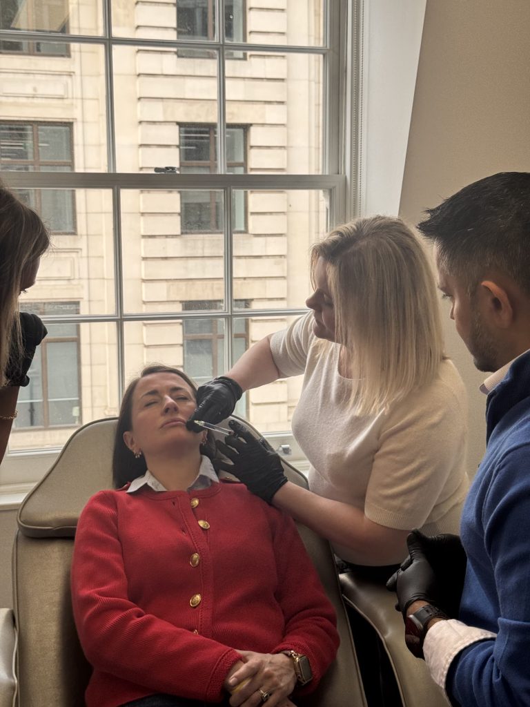 A delegate injects a model patient's lower face while two colleagues observe during supervised training.